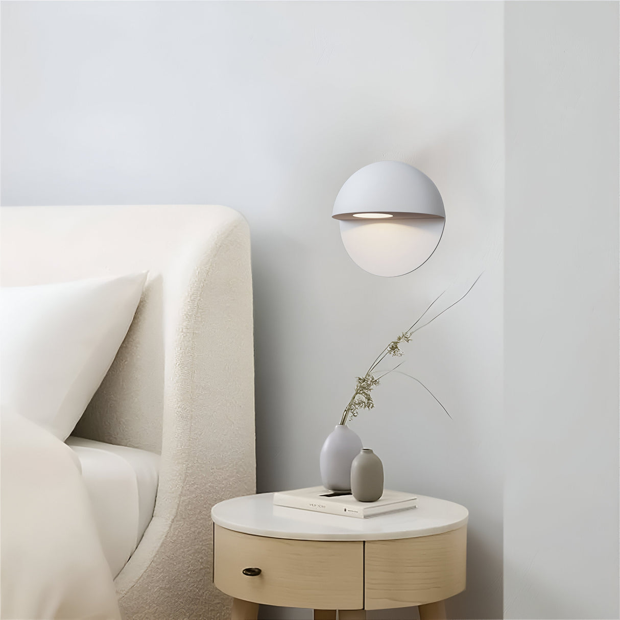 Minimalist bedroom features a white, curved headboard and a Mezzo Outdoor LED Wall Light 3000K - White. A durable aluminum side table supports a gray vase with dried flowers, books, and warm white lighting to enhance the serene, modern aesthetic.