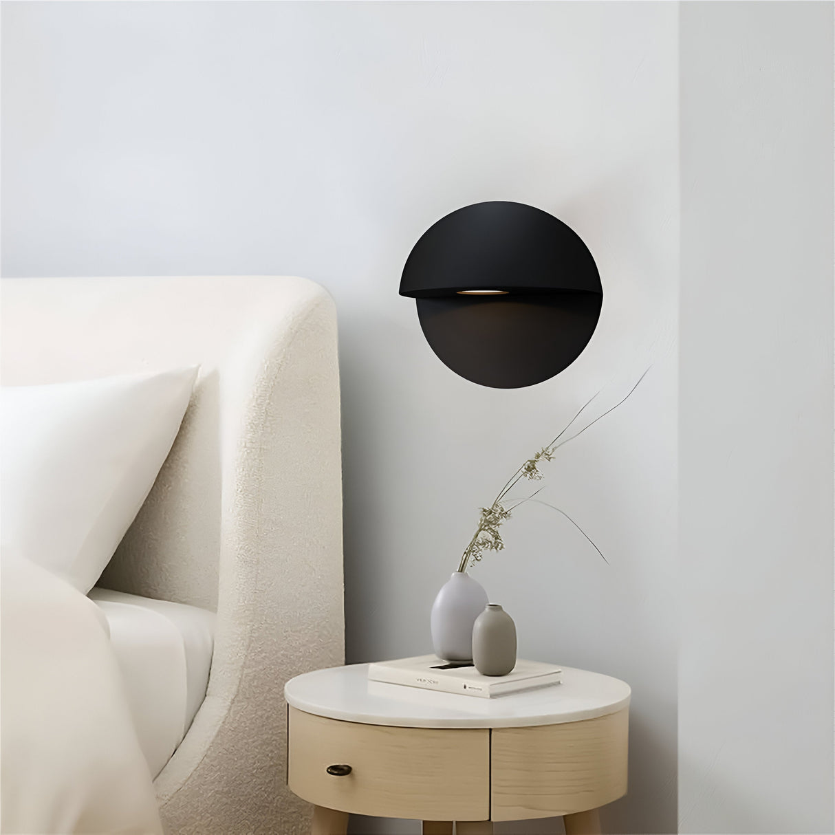 A minimalist bedroom includes a white, textured headboard, a corrosion-resistant Mezzo Outdoor LED Wall Light in black that emits a warm white glow. A beige nightstand is adorned with a small vase of dried plants and a closed book, creating simple and modern decor.