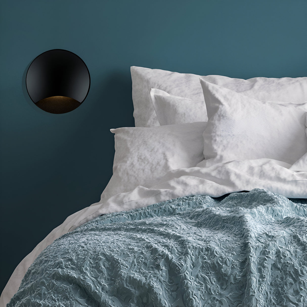 A neatly made bed with white pillows and sheets is complemented by a textured light blue blanket. The teal wall behind features the Biscotti Outdoor 1 Beam LED Recessed Wall Light in black on the left, casting a subtle glow to enhance the serene ambiance.