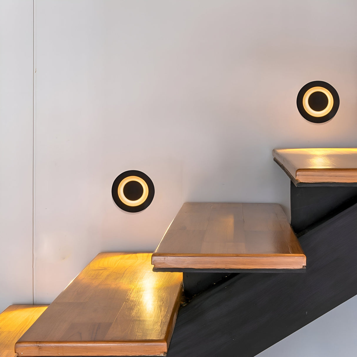 A modern staircase with wooden steps and a black frame is lit by two Limo Outdoor LED Recessed Wall Lights - Black. Mounted on the white wall beside the steps, these energy-efficient lights cast a warm glow, enhancing the ambiance.