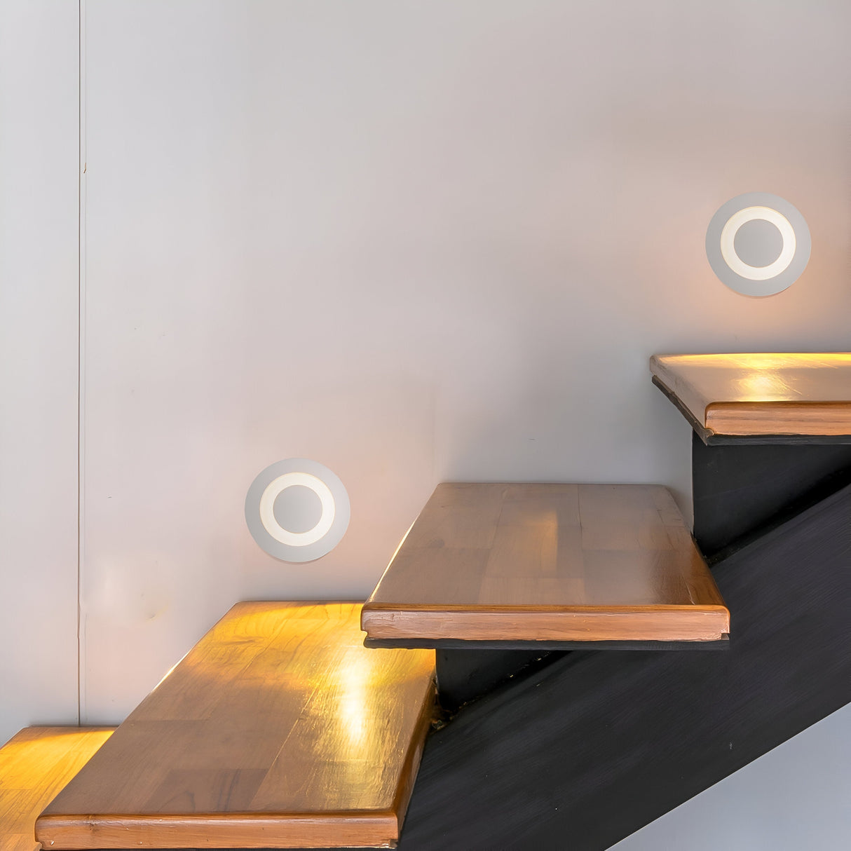 A modern staircase with wooden steps and a black frame is lit by the Limo Outdoor LED Recessed Wall Light - White. These lights cast a warm glow on the steps and white wall, creating a minimalist and contemporary atmosphere.