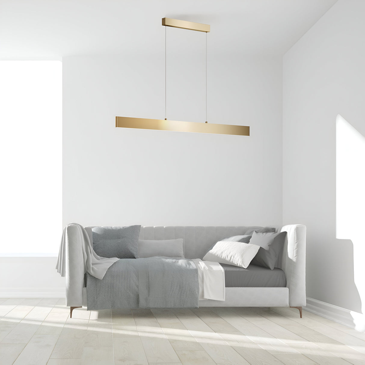 A minimalist living room showcases a light gray sofa adorned with blankets and cushions against a white wall. Above, the Step LED Long Linear Suspension Light 3000K 30W in Gold adds elegance. Sunlight filters through a large window, illuminating the light wood flooring.