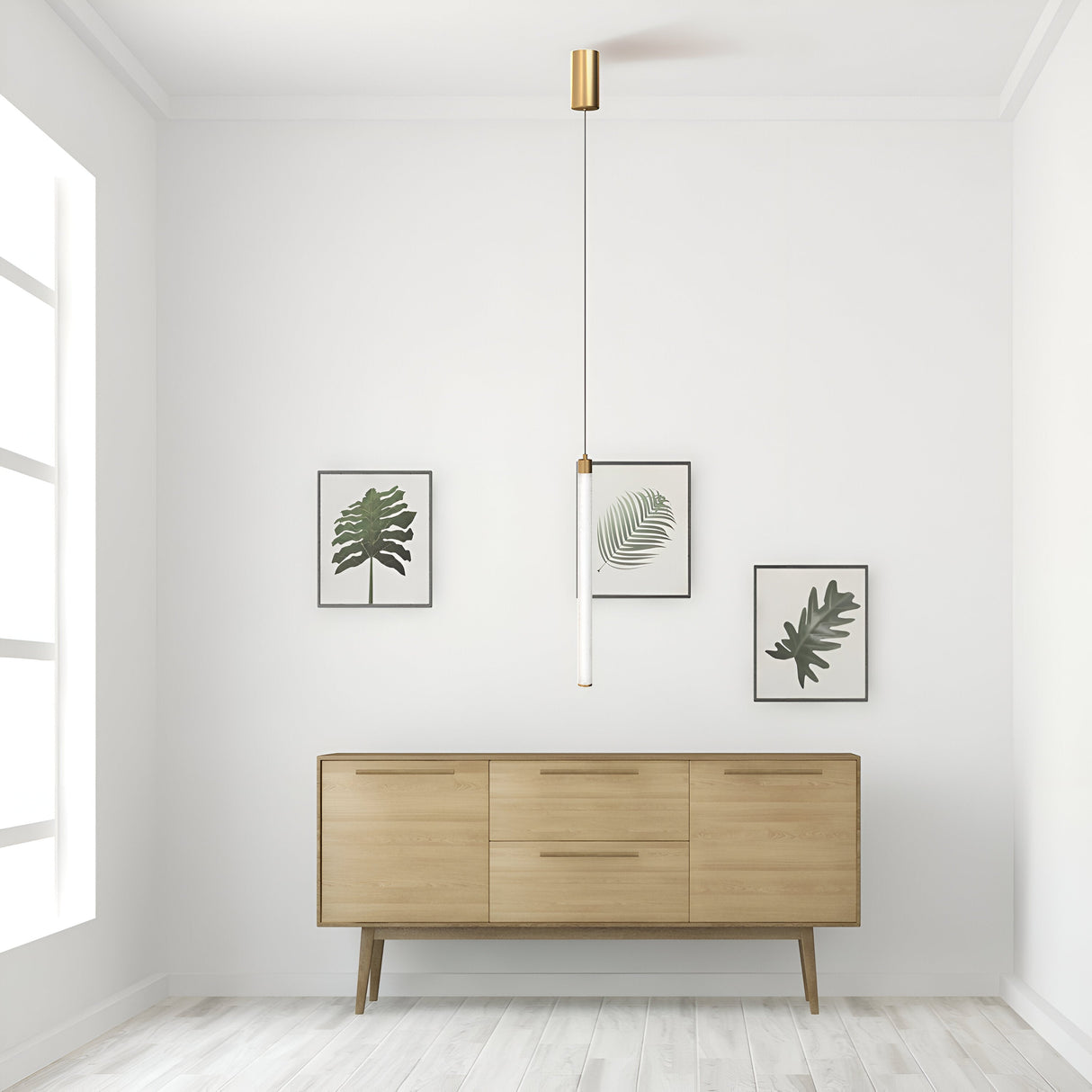 A minimalist room showcases a light wood sideboard against a white wall. Above it, three framed botanical prints are displayed. The Ray LED Long Pendant Light in gold and white hangs elegantly from the ceiling. Natural light floods through a tall window on the left.