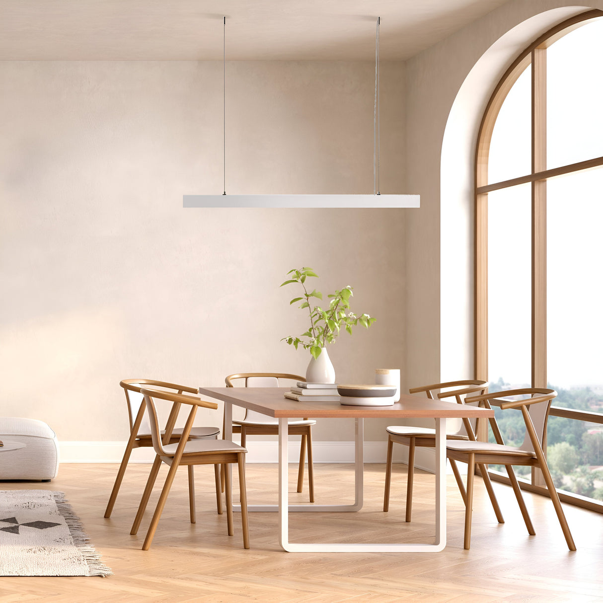 A minimalist dining room showcases a wooden table with four matching chairs adorned with a white vase of green leaves. Large arched windows on the right illuminate beige walls and a light wood floor, while the BASIS LED Linear Suspension Light - White enhances the modern aesthetic overhead.