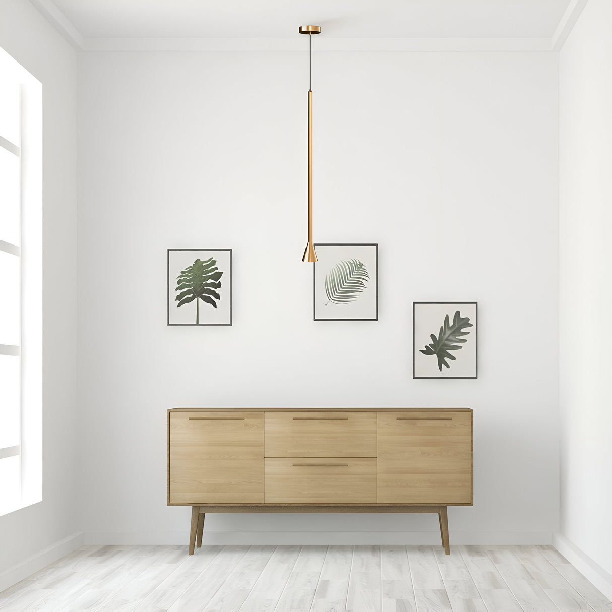 A minimalist room has a wooden sideboard against a white wall, with three framed leaf illustrations above. The Arrow 1 Light Long Cone Pendant - Gold hangs gracefully from the ceiling. Light wood flooring perfectly complements the neutral decor.