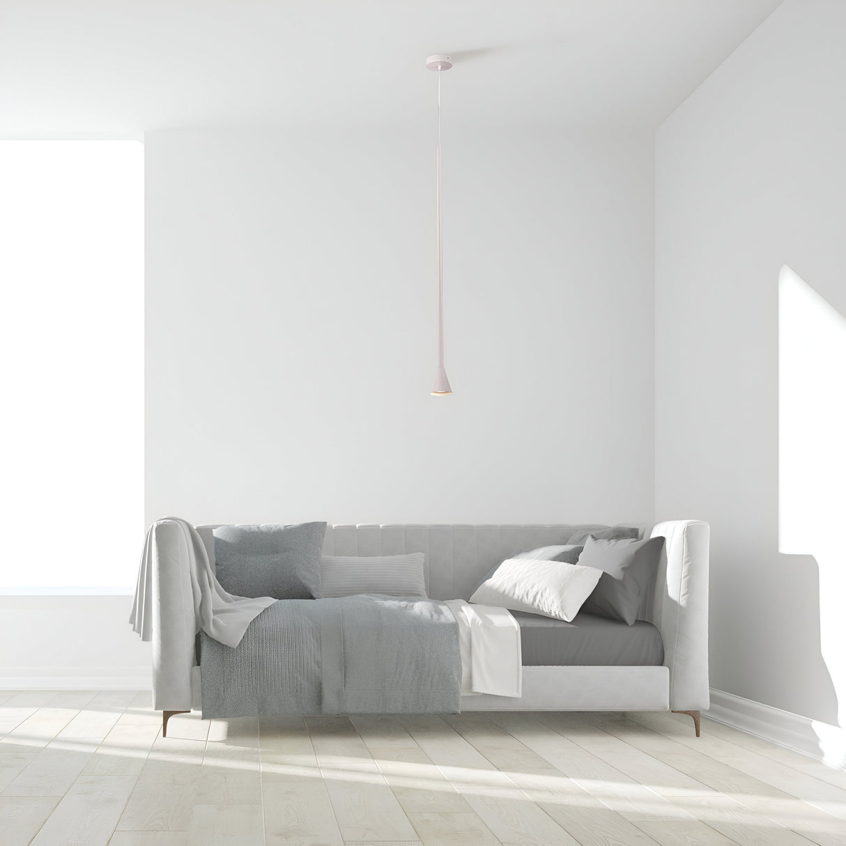 A minimalist living room features a light gray sofa adorned with a quilt and cushions against a white wall. Natural light pours through large windows, illuminating the wooden floor and emphasizing the sleek, modern design of the Arrow 1 Light Long Cone Pendant - White.