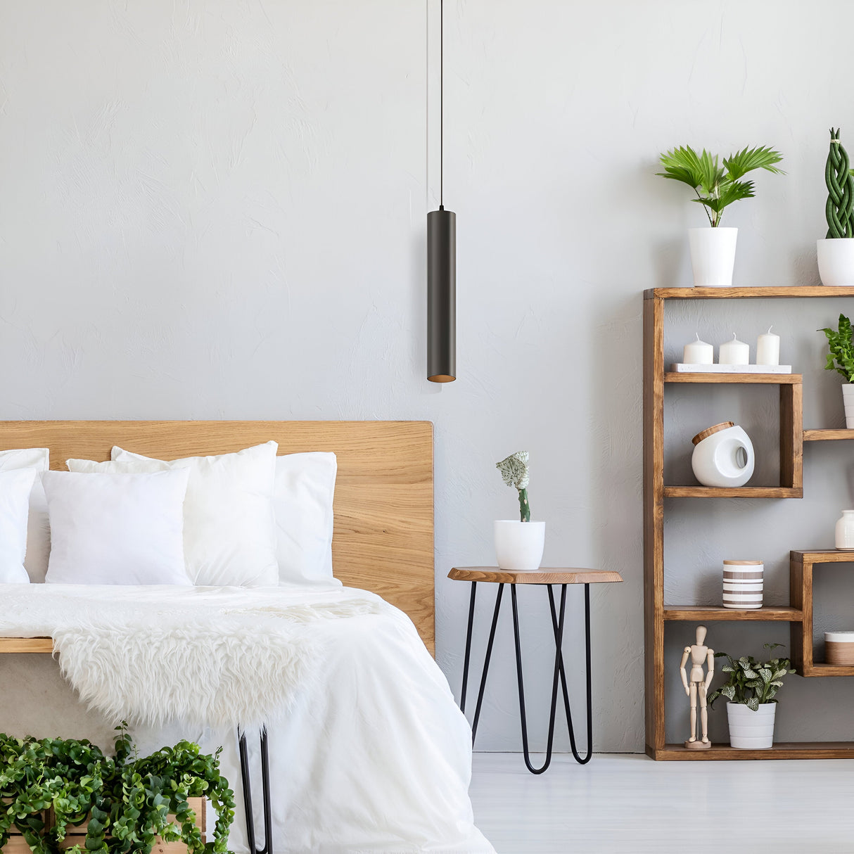 In a minimalist bedroom, a wooden bed frame with white bedding and a sheepskin throw sits beside a small round table featuring a plant. A geometric wooden shelf holds plants and decor, while energy-efficient lighting is provided by the Focus LED Pendant Light 3000K - Black above.