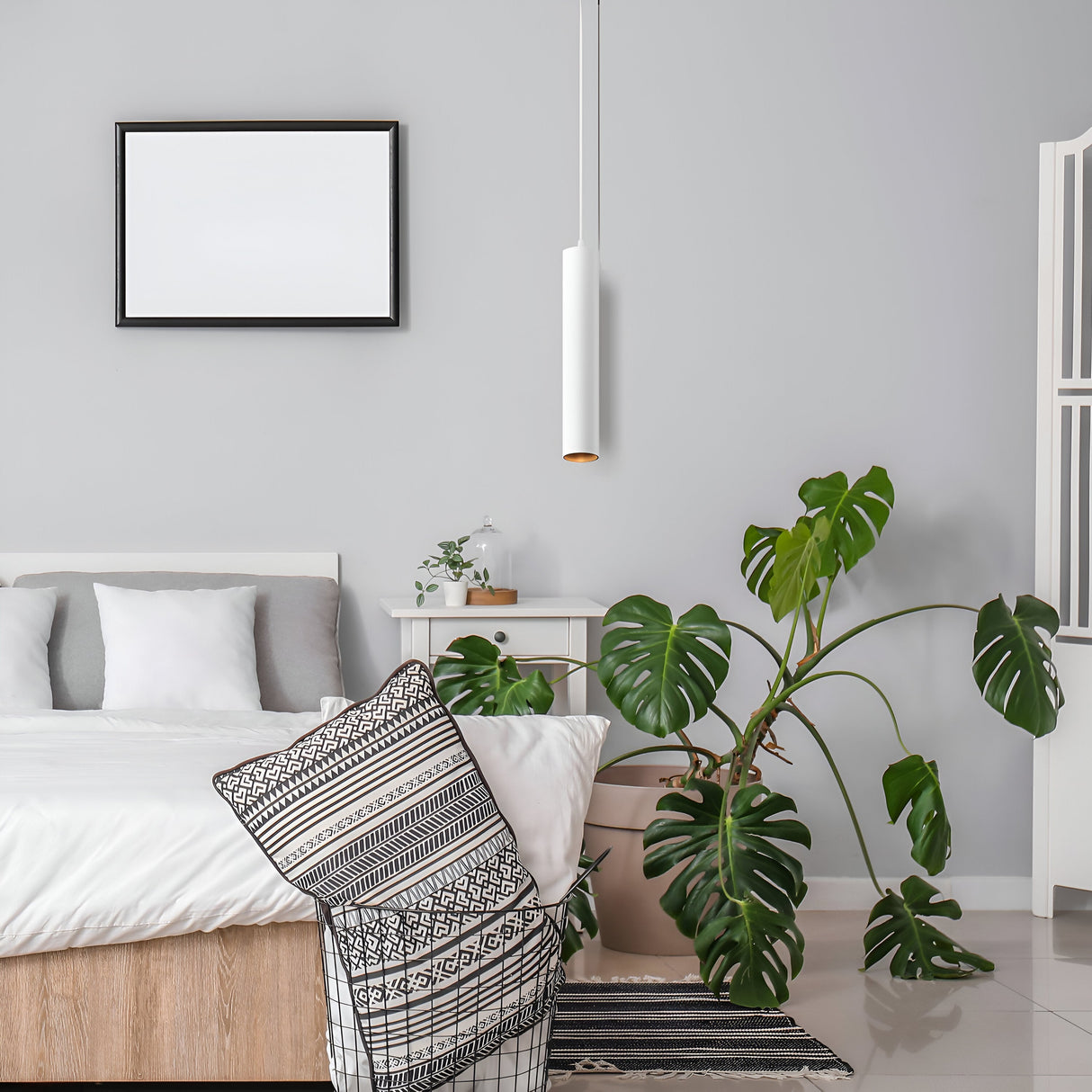 A modern bedroom features a wooden bed with white bedding and patterned pillows. A large monstera plant stands beside a nightstand with a white lamp. The Focus LED Pendant Light 3000K in white enhances the minimalistic decor, while a blank picture frame adorns the light gray wall.