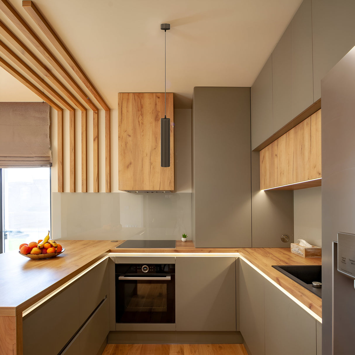 A modern kitchen features a U-shaped wooden counter, built-in oven, and sleek grey cabinets with a black metal finish. A fruit bowl rests beneath the Focus Single Pendant Light - Black, while a small window brightens the rooms minimalist design and industrial aesthetic.