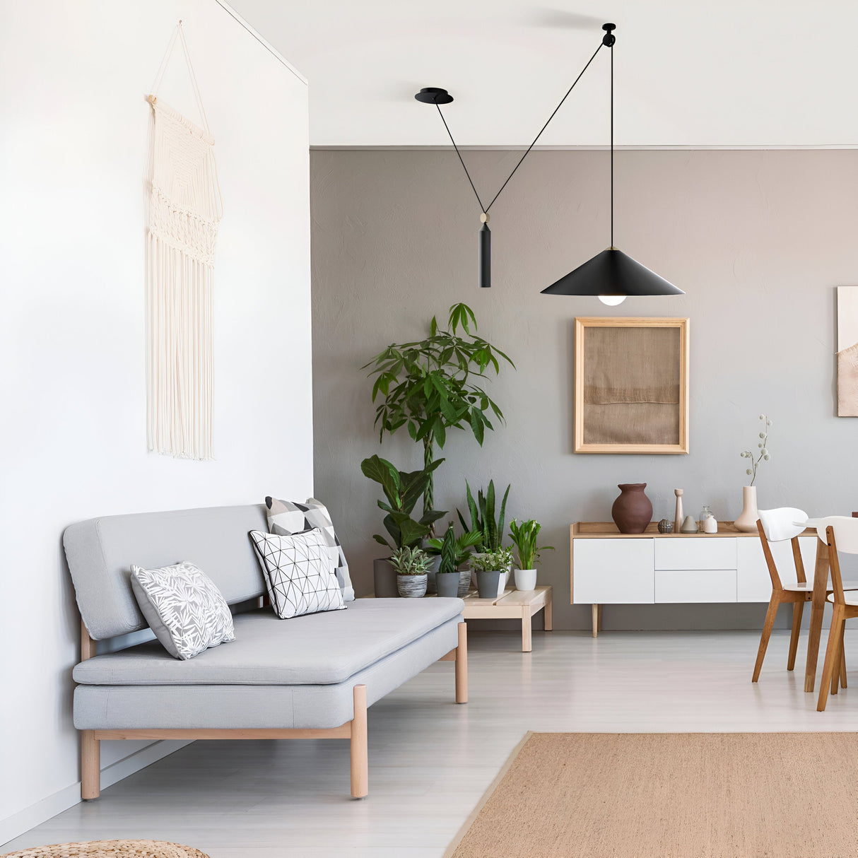A minimalist living room features a light gray sofa with patterned cushions under the Peso Adjustable Rise & Fall Conical Pendant Light in black. A macramé wall hanging, a potted plant, and an adjustable-height sideboard displaying decor near a dining table with chairs complete the scene.