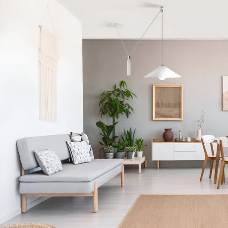 A modern aesthetic graces the living room with a light gray sofa adorned with patterned cushions. Potted plants complement the white sideboard and macrame wall hanging. The dining table features chairs beneath the stylish Peso Adjustable Rise & Fall Conical Pendant Light in white.