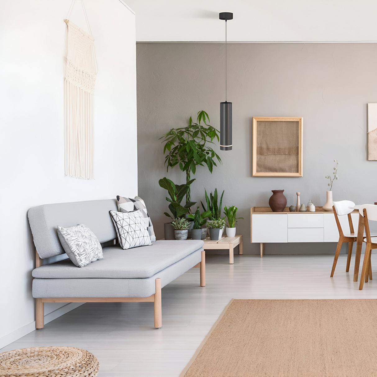 A modern living room features a light gray sofa with patterned cushions, surrounded by various potted plants. A white sideboard with decor and a dining table with wooden chairs complement the light gray walls, all elegantly illuminated by the Orlo LED Pendant Light 3000K - Black above.