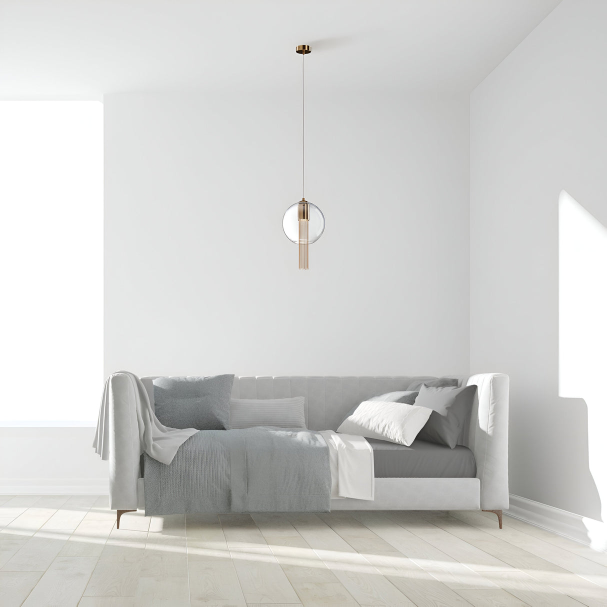 A minimalist living room showcases a modern gray sofa adorned with a blanket and cushions. A Flusso Long Chain Pendant Light - Bronze With Clear Glass Ball hangs elegantly above, as sunlight streams through a large window, illuminating the white walls and wooden floor.