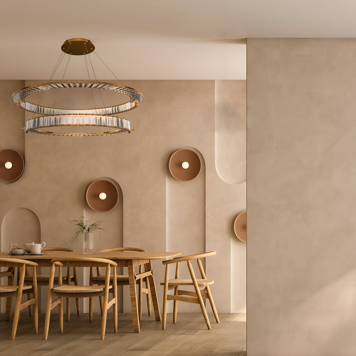 A minimalist dining area features a wooden table and chairs beneath the Krone 2 Tier LED Circular Crystal Suspension Light - Gold, adding modern luxury. Round fixtures adorn the textured beige wall, while a small vase with flowers enhances the serene elegance.