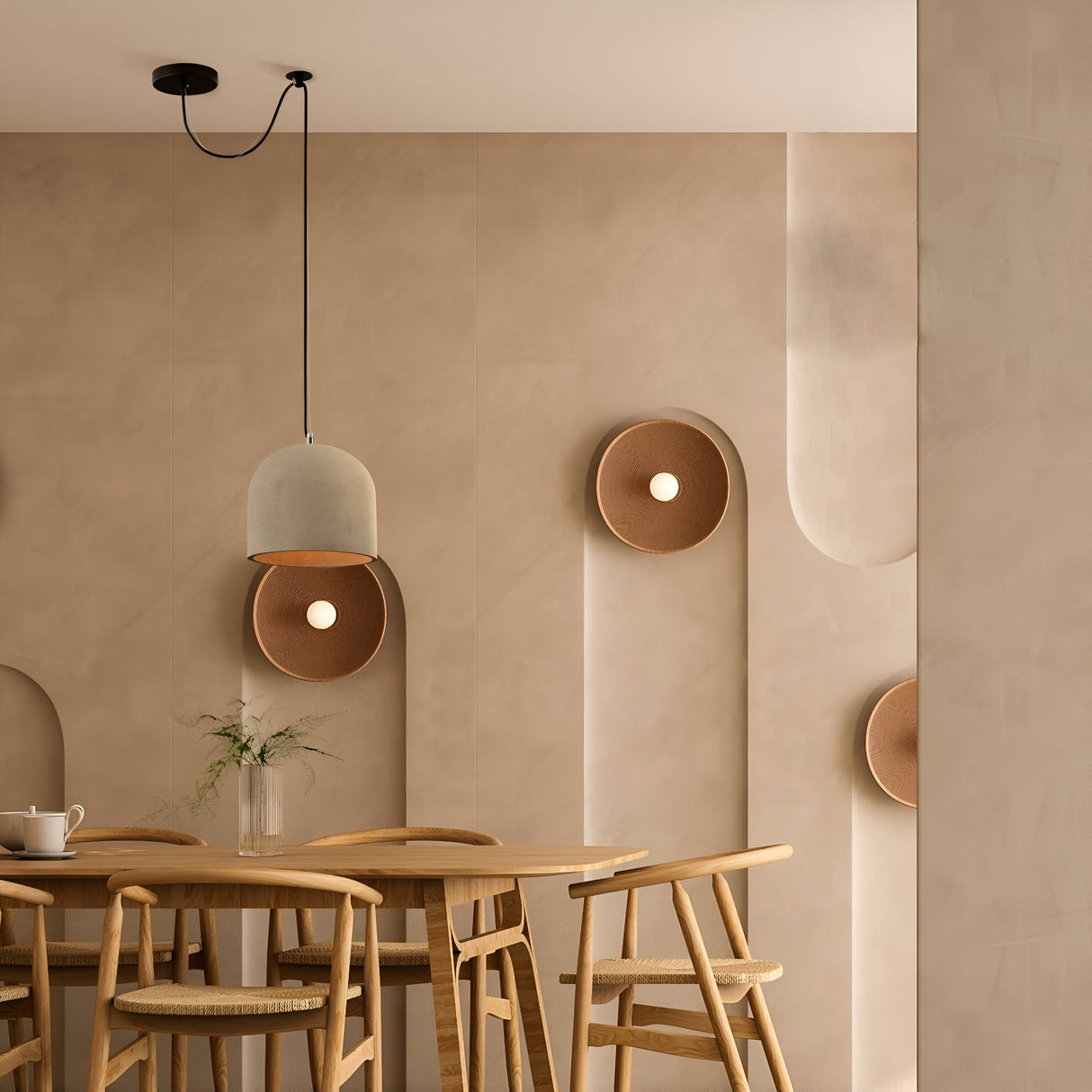 A minimalist dining area features a wooden table and chairs, illuminated by the Broni Pendant Light 20cm in black with a grey concrete shade. Wall-mounted circular lights complement the textured neutral-toned walls, while a small vase of flowers adds to the decor.