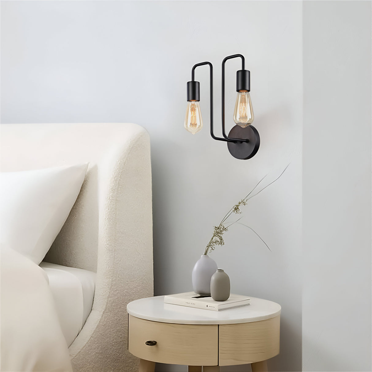 A minimalist bedroom scene showcases a beige upholstered bed and round wooden nightstand. The nightstand holds two books and a small vase with dried flowers, while the Gilbert 2 Light Wall Light in black with an open bulb design casts a soft glow from above.