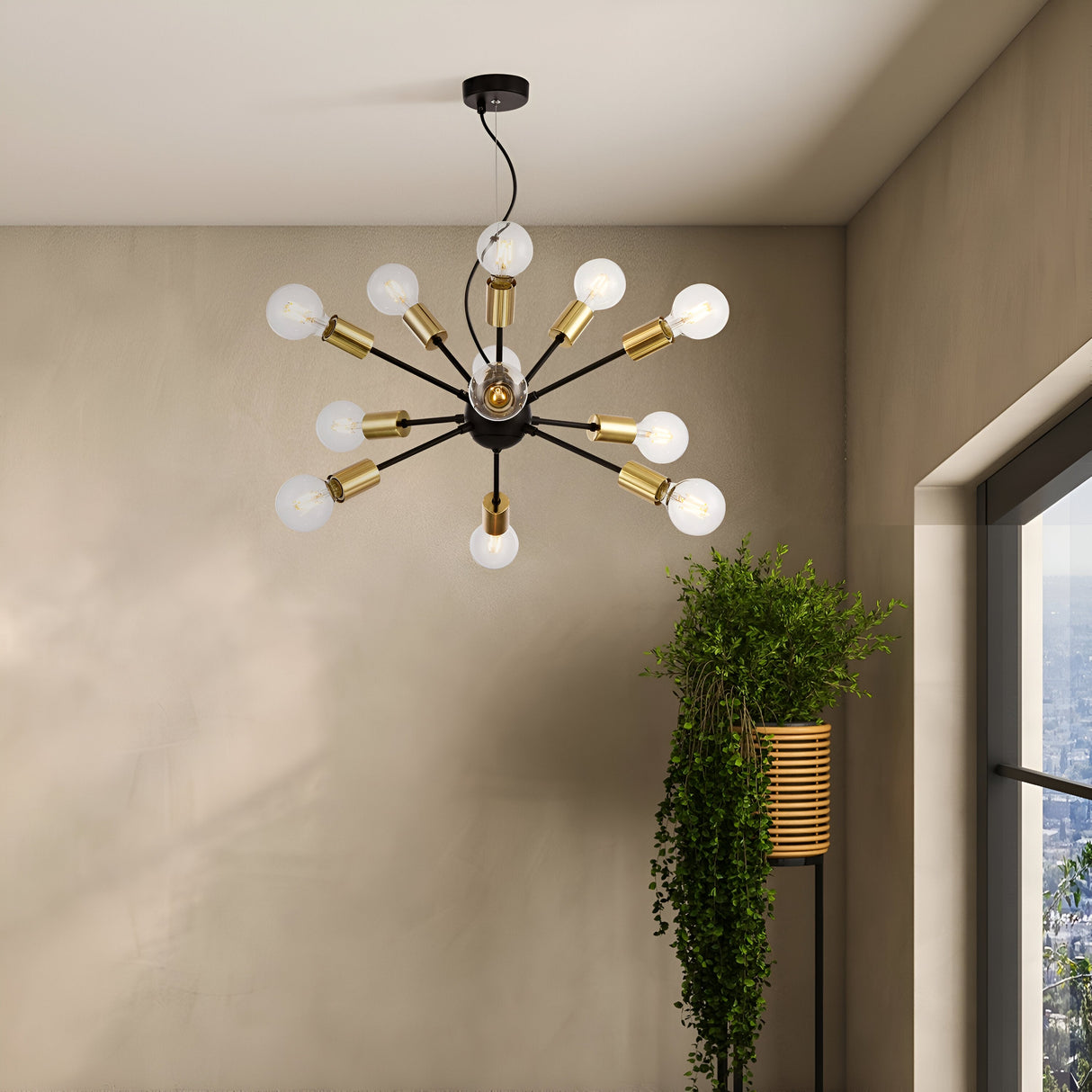 The Jackson 12 Light Chandelier - Black, with multiple extending arms each ending in a round bulb, hangs from the ceiling. A lush plant in a tall woven basket stands by a window, partially revealing a cityscape backdrop.