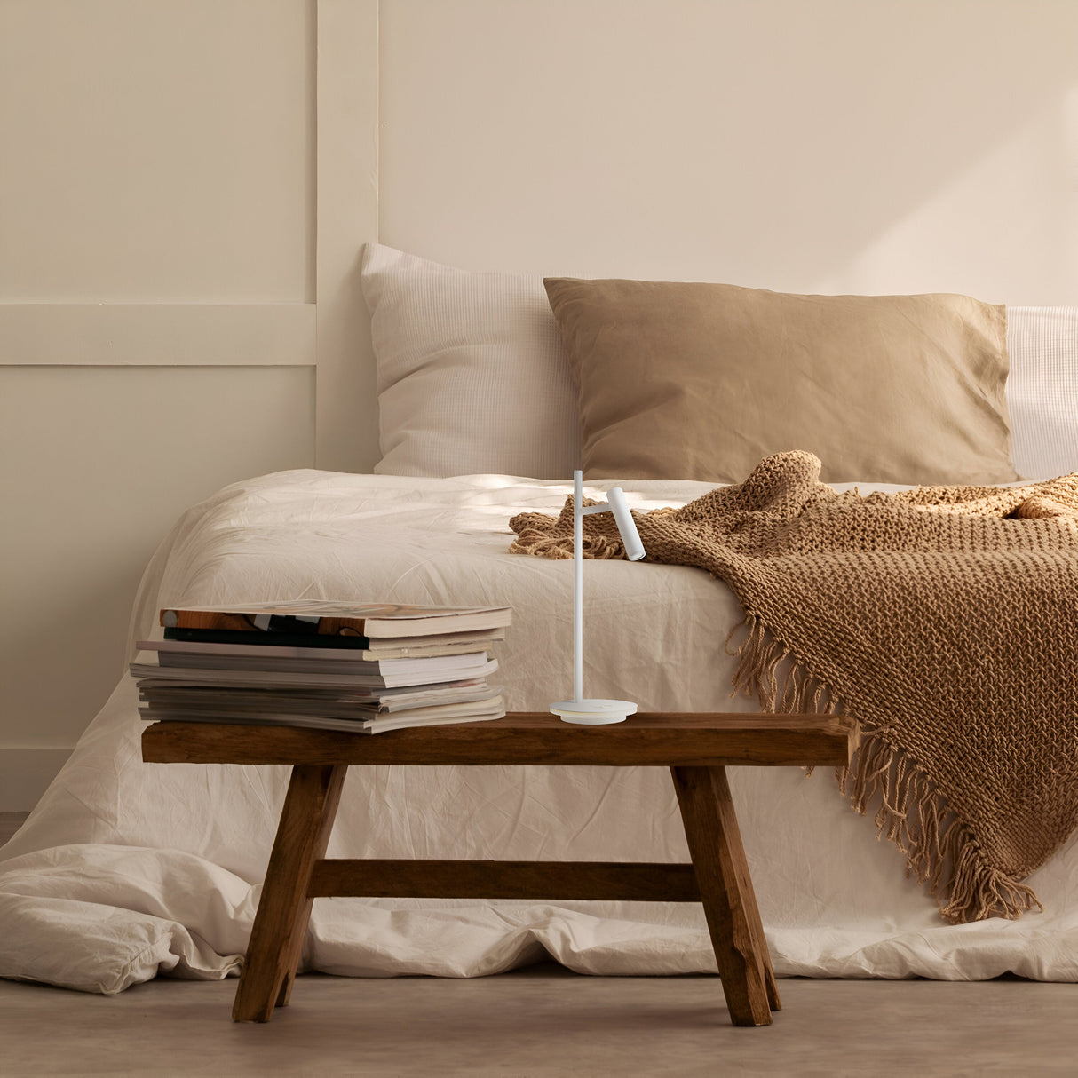 In a cozy bedroom, a neatly made bed with beige and white bedding is complemented by a wooden bench serving as a side table. The Estudo LED Table Lamp - White sits on it alongside books. A warm brown knit blanket is draped over the bed, enhancing the lighting ambiance.