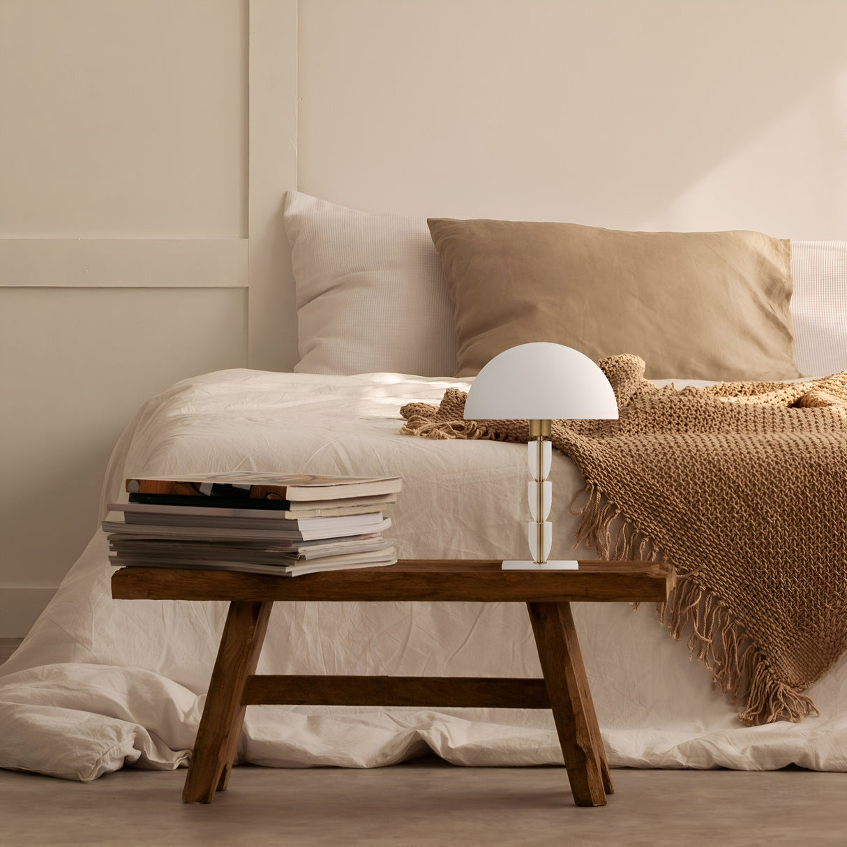 A cozy bedroom setup features a bed with white and beige bedding, a beige throw, and pillows. A wooden bedside table holds a stack of books and the Prima Table Lamp in brass with a white shade. The rooms warm, neutral color scheme is complemented by subtle brass frame accents.