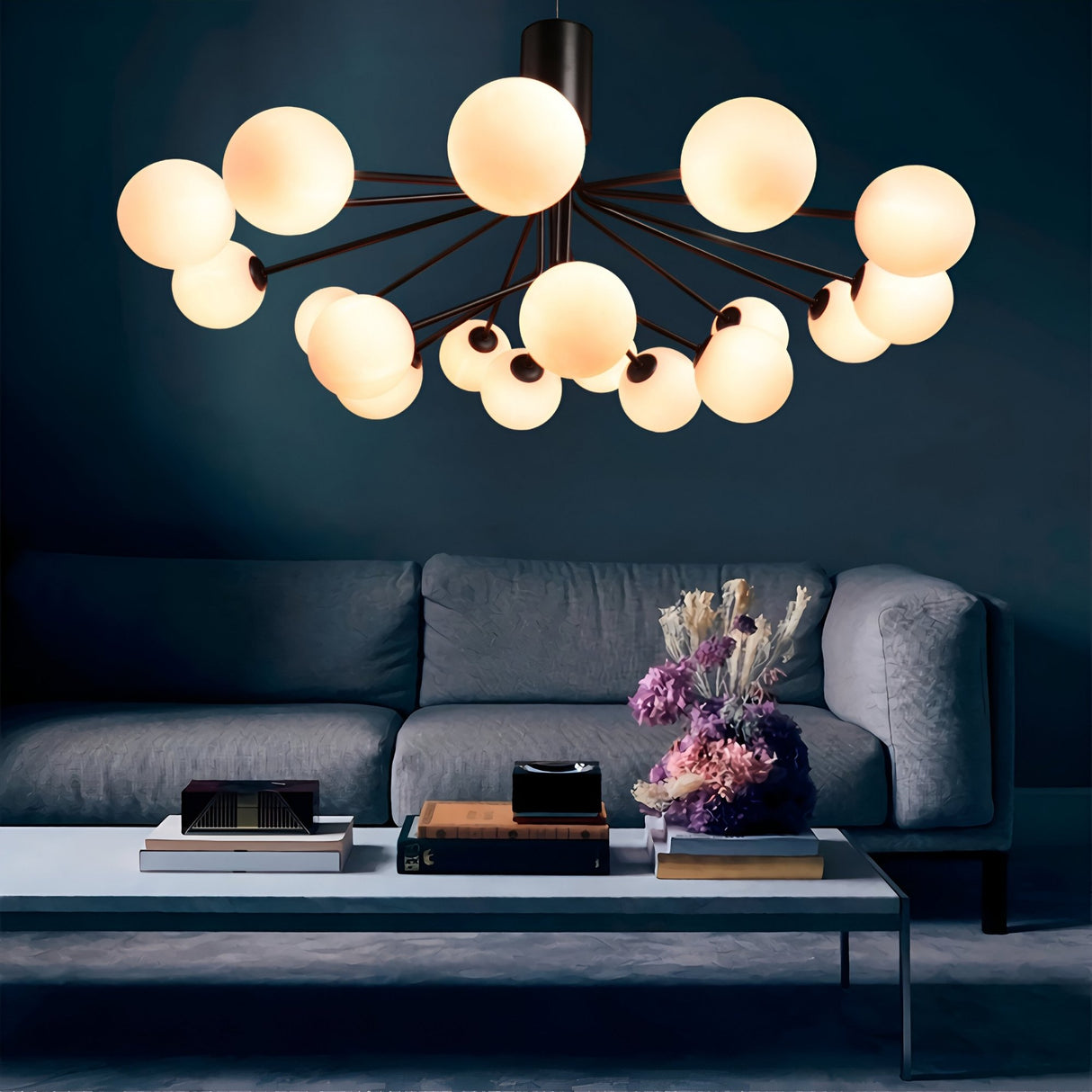 A modern living room with a grey sofa, white coffee table topped with books and flowers, and the Ivy 18 Light Chandelier - Black showcasing sleek metal arms and glowing opal glass shades overhead.