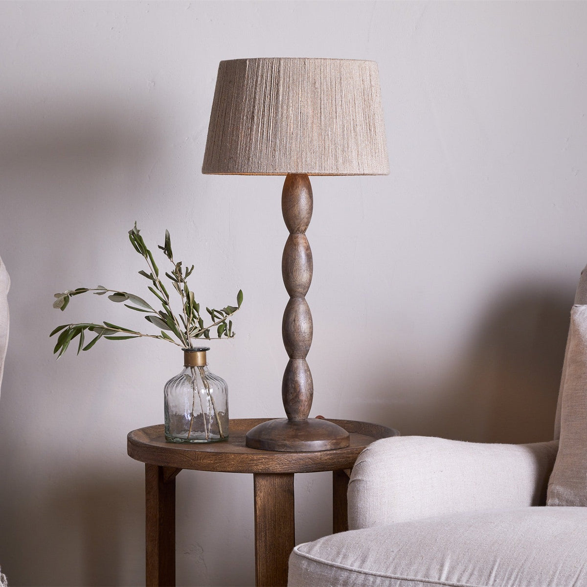 Konali Mango Wood Table Lamp Base in ash stain with beaded spindle column, round foot and tapered linen shade, shown on a wooden side table beside a glass vase.