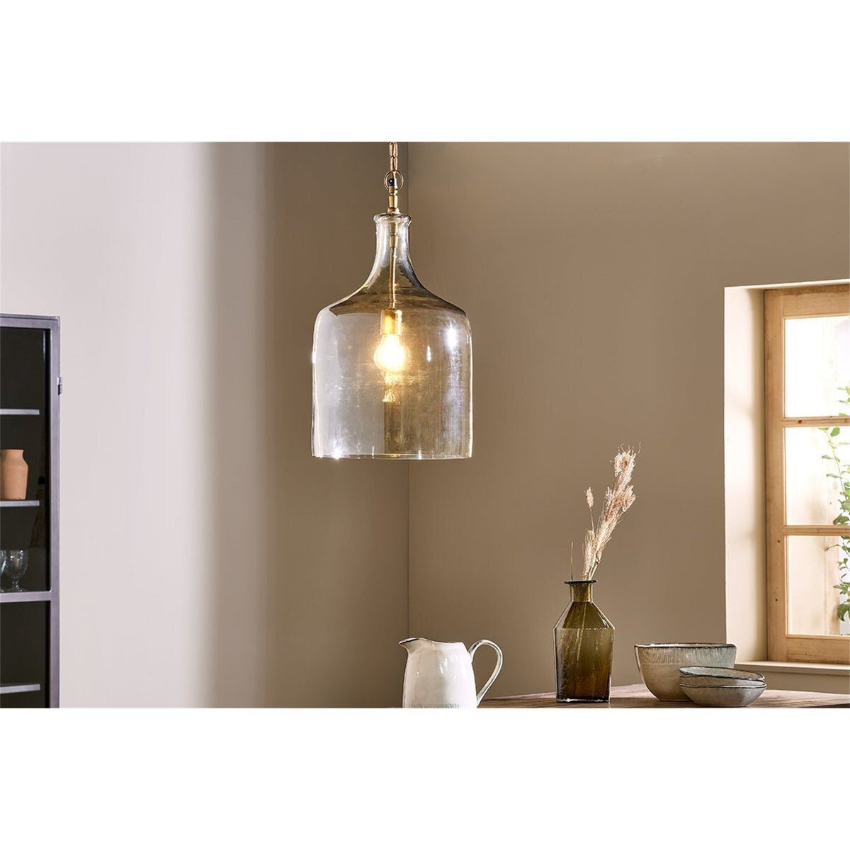 Kalsi Large Recycled Glass Pendant Light in Lustre finish, a tall jar-shaped shade with brass collar and visible lampholder, shown lit above a dining table in a warm-toned room.