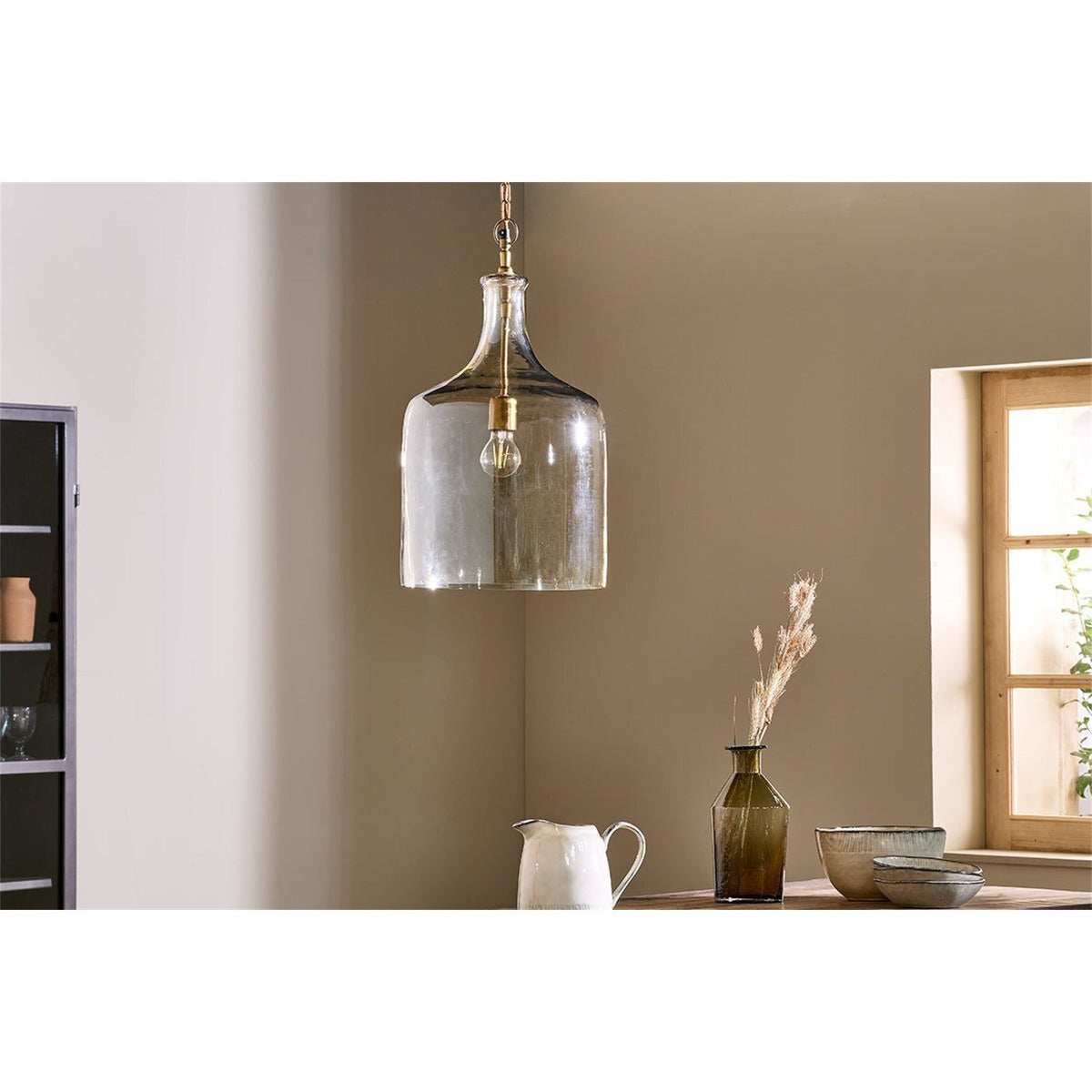 Kalsi Large Recycled Glass Pendant Light in Lustre finish, a tall flagon-shaped glass shade with brass collar and visible lampholder, hung in a dining area with a wooden table and decor nearby.