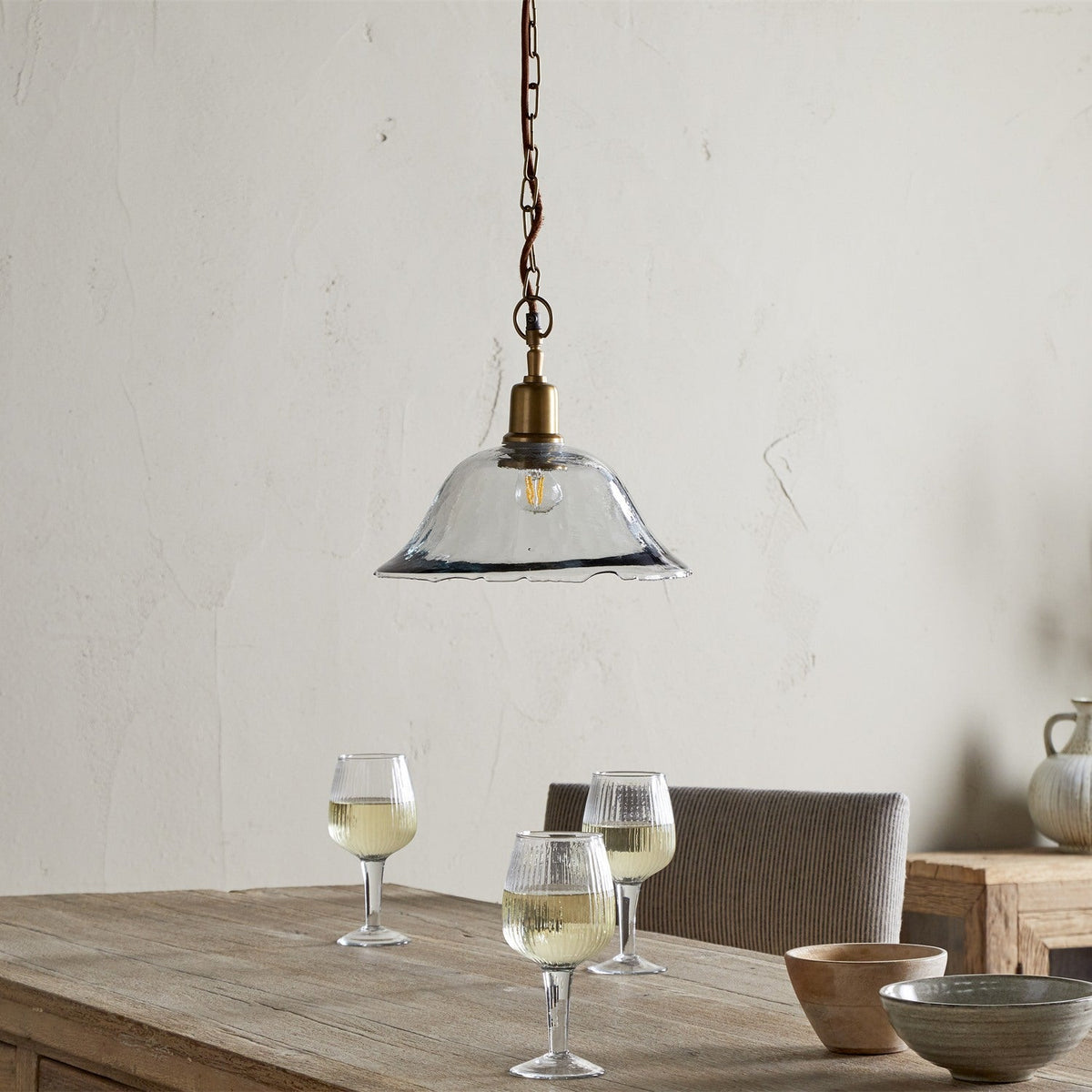 Keju Recycled Glass Pendant Light in Clear, a wide shallow bell shade with rippled rim on a brass stem and chain, hanging above a wooden dining table.