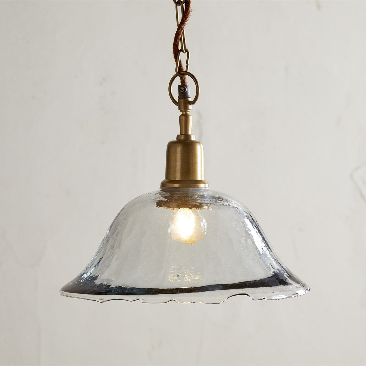 Keju Recycled Glass Pendant Light in Clear with a rippled, bubbled bell-shaped shade, brass collar and chain, and rust fabric flex, shown on a plain light background.