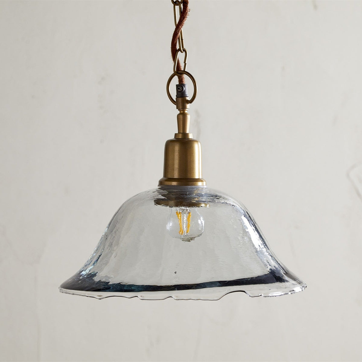 Keju Recycled Glass Pendant Light in Clear, a wide shallow bell-shaped shade with a rippled rim on a brass collar and stem, hung from a brass chain with rust fabric flex.