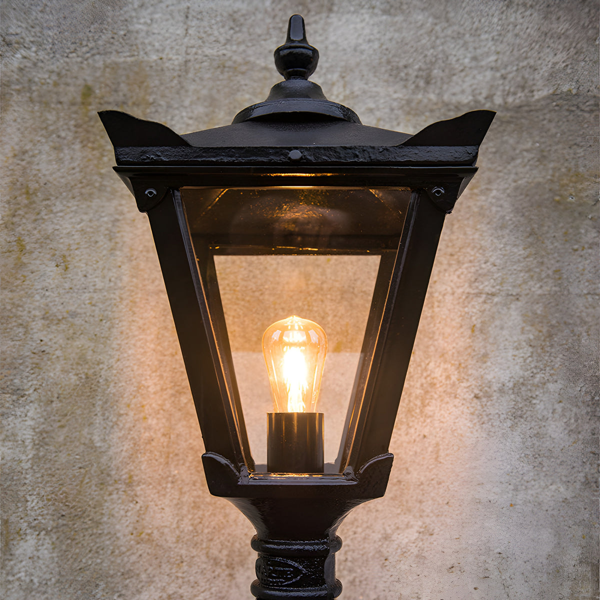 Victorian-style black cast iron replacement lantern head with glass panels and lit bulb, ornate finial top, shown against a weathered stone wall.