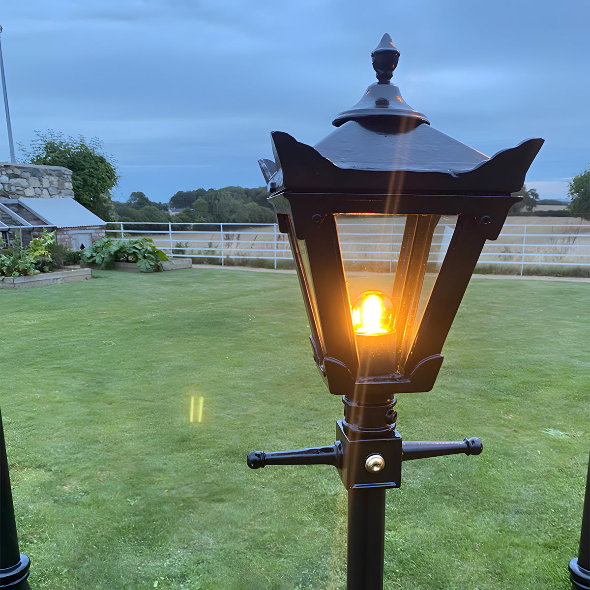Victorian cast iron replacement lantern head in black, lit and mounted on a post in a garden setting with lawn and countryside visible behind.