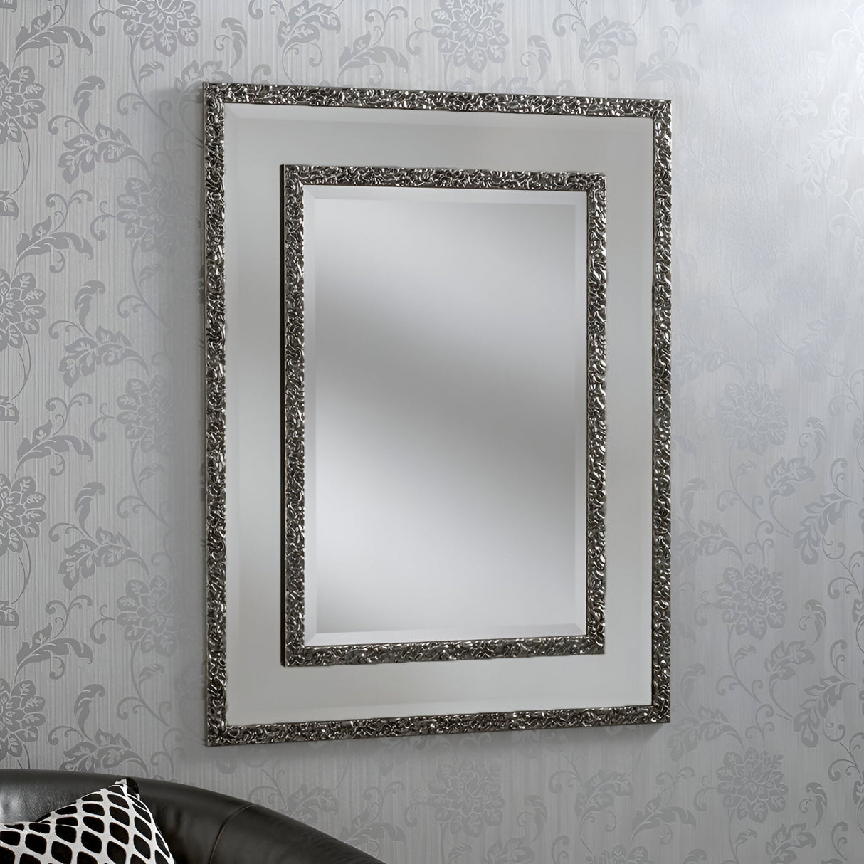 The Lava Rectangle Wall Mirror Grey, measuring 108x82cm, with an ornate grey frame, hangs on a wall covered in patterned wallpaper. At the bottom left corner, a glimpse of a black leather couch adorned with a black and white patterned pillow is visible, enhancing the modern decor.