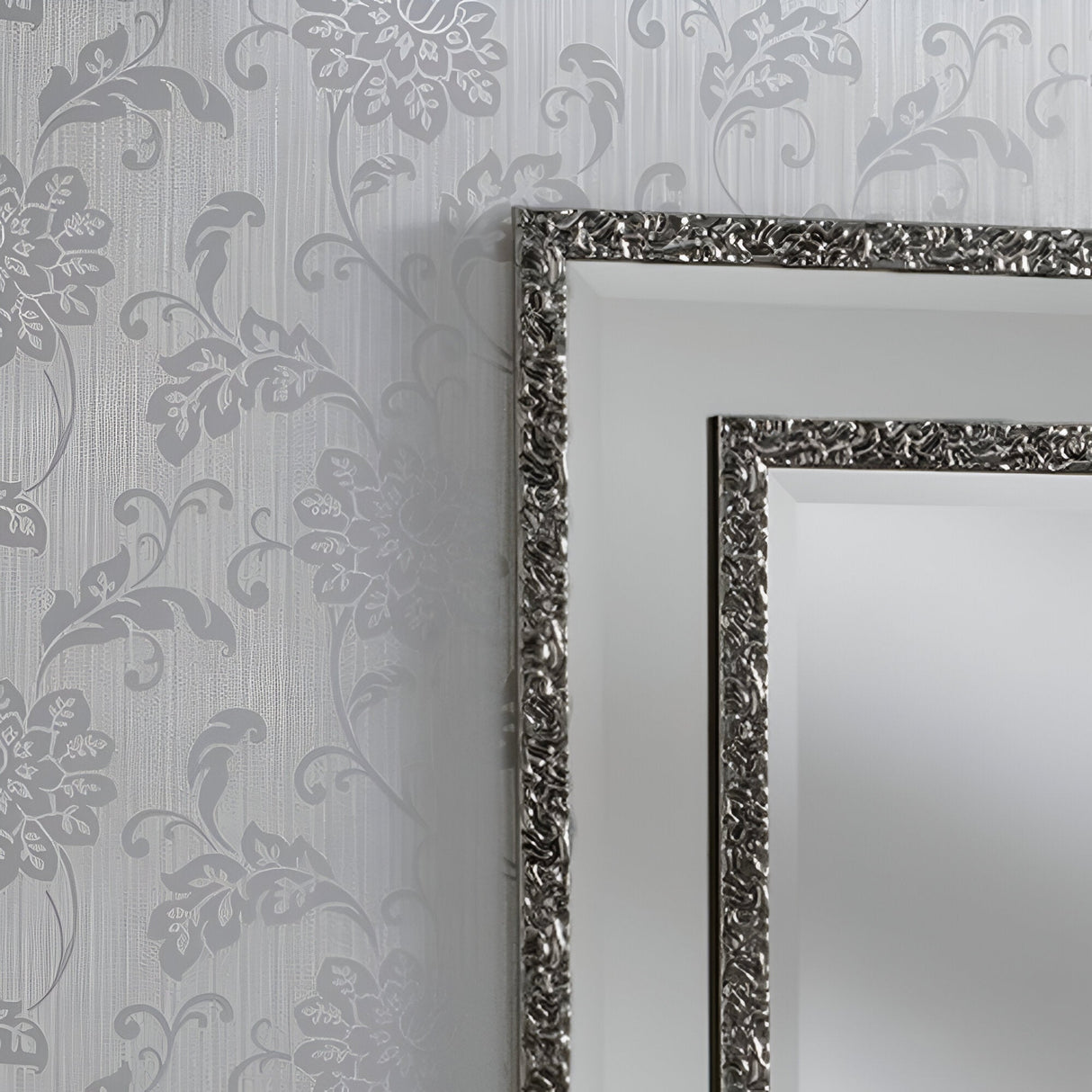 A Lava Rectangle Wall Mirror in Grey, measuring 108x82cm, is mounted on a wall featuring floral-patterned wallpaper in shades of gray. The ornate design of the mirror's frame pairs beautifully with the intricate wallpaper, creating an elegant and cohesive look that seamlessly integrates modern decor into the space.