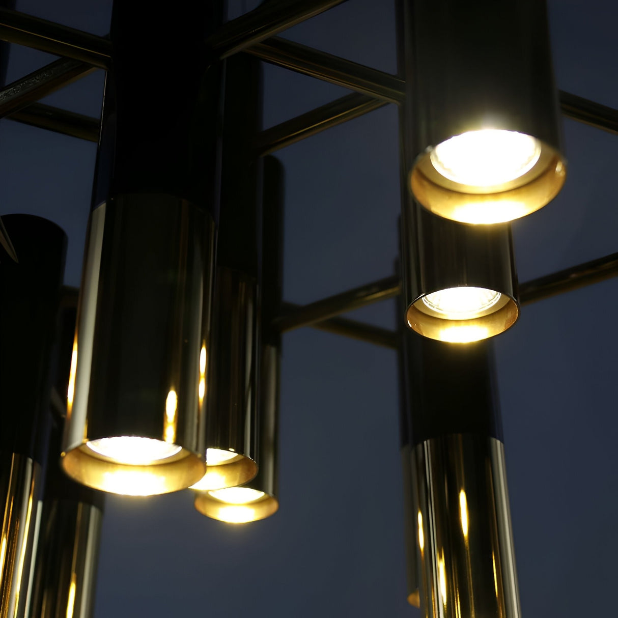 Close-up of the Leonard 19 Light LED Chandelier in black and gold, featuring illuminated cylindrical pendants with metallic finishes, highlighting energy-efficient lighting and a sleek modern design against a dark background.