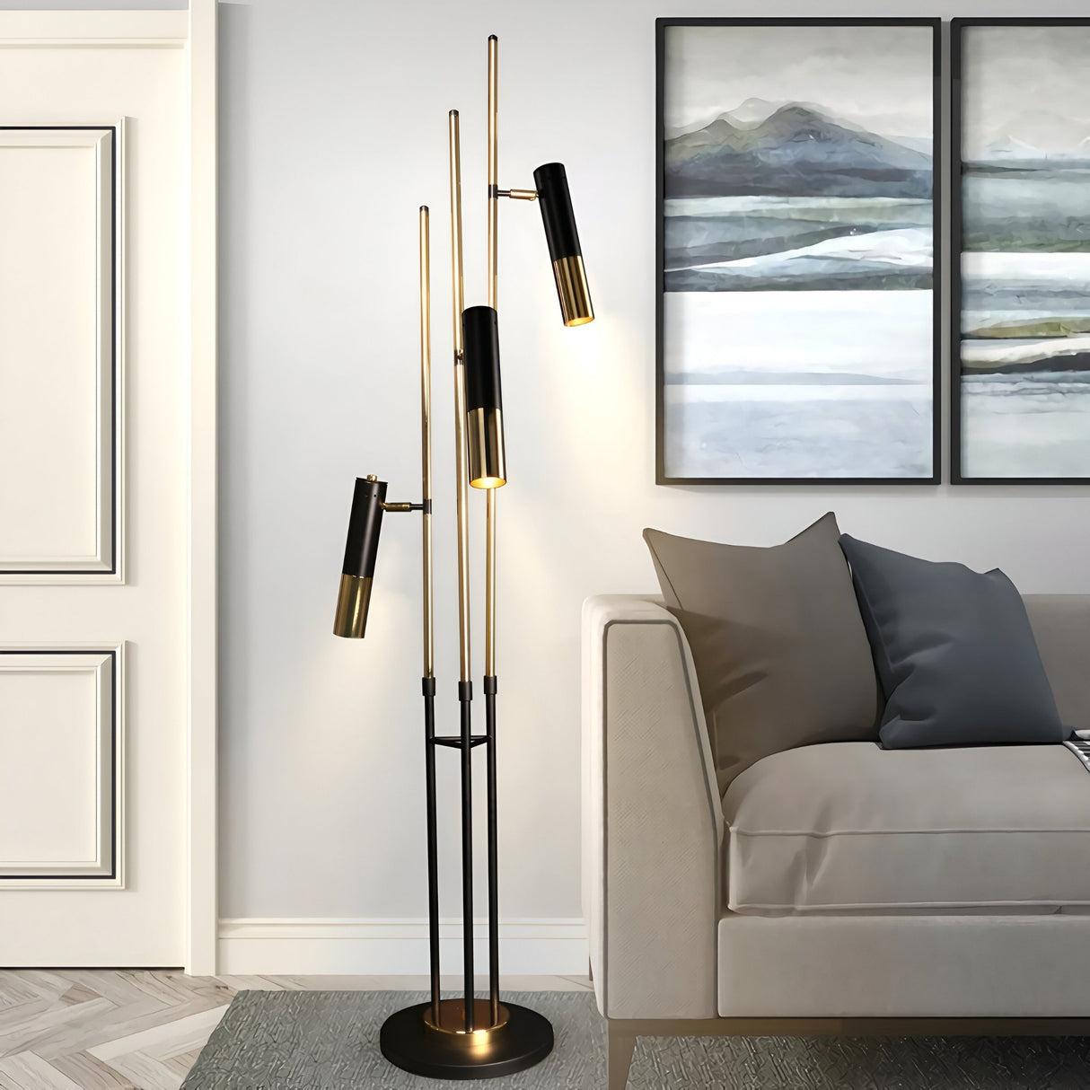 A modern living room with a beige sofa, gray pillows, the Leonard 3 LED Floor Lamp in black & gold for energy-efficient lighting, painted landscape wall art, and a white paneled door in the background.