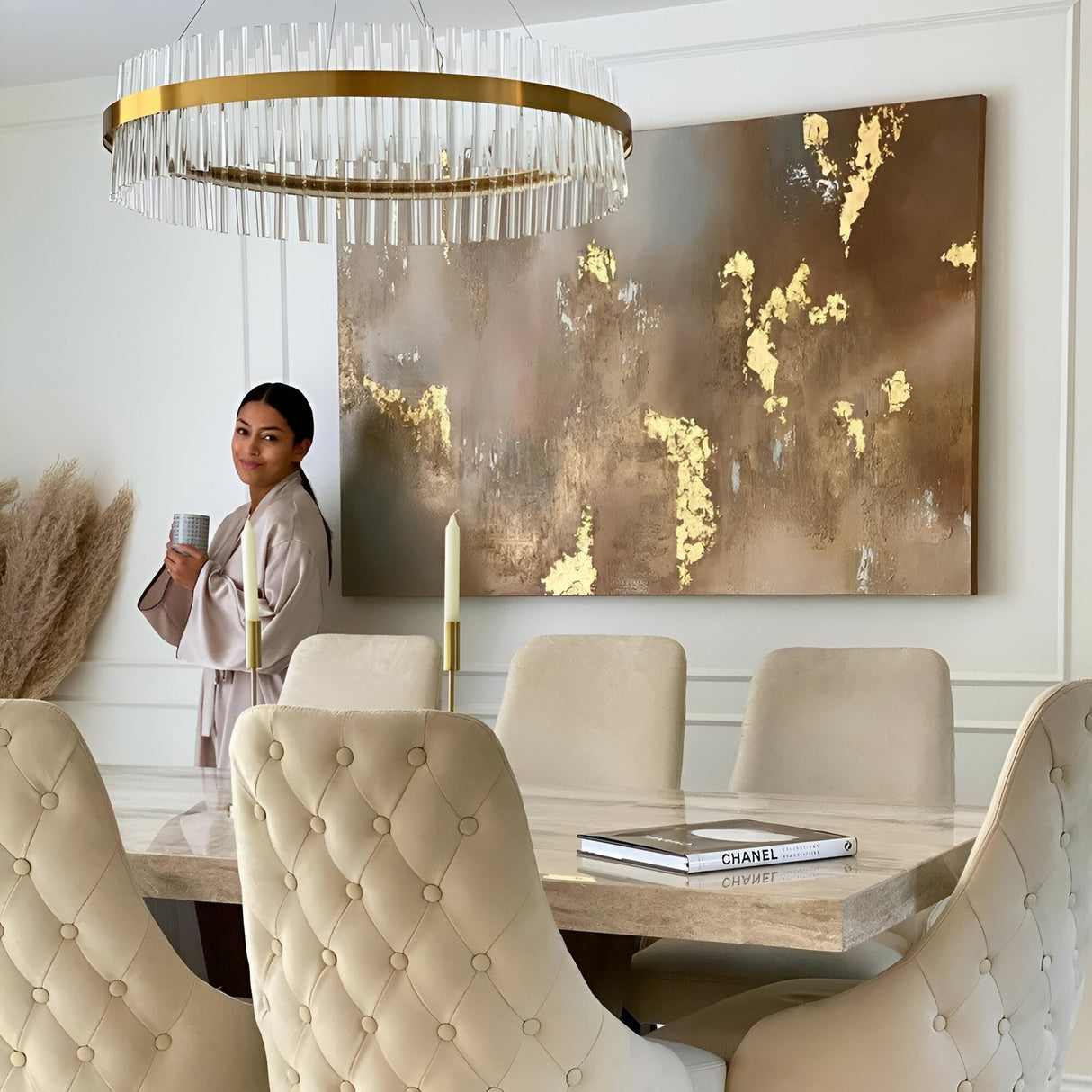 A woman in a light robe holds a mug by a modern dining table with beige tufted chairs, candles, books, and abstract gold-accented wall art beneath the Liberty LED Crystal Glass Chandelier - Gold in a stylish, neutral-toned room.