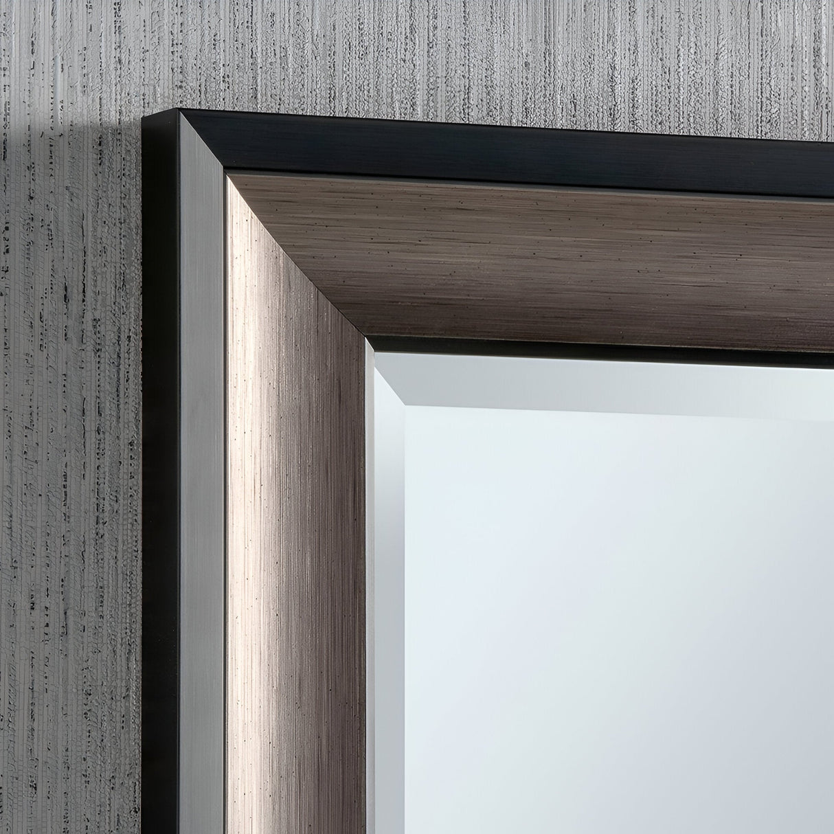Close-up of the Liberty Rectangle Wall Mirror - Black & Brushed Grey mounted against a textured gray wall. Its sleek, metallic frame showcases straight lines with a brushed finish in black and grey. The mirror reflects soft, ambient light, enhancing its clean and sophisticated appearance with modern design.