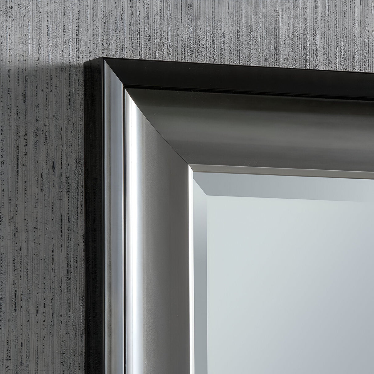 Close-up of the Liberty Rectangle Wall Mirror - Gloss Black & Gunmetal Grey Silver corner against a textured gray wall. The mirror's metal frame exudes modern elegance with its sleek, reflective design, making it a versatile addition to any space.