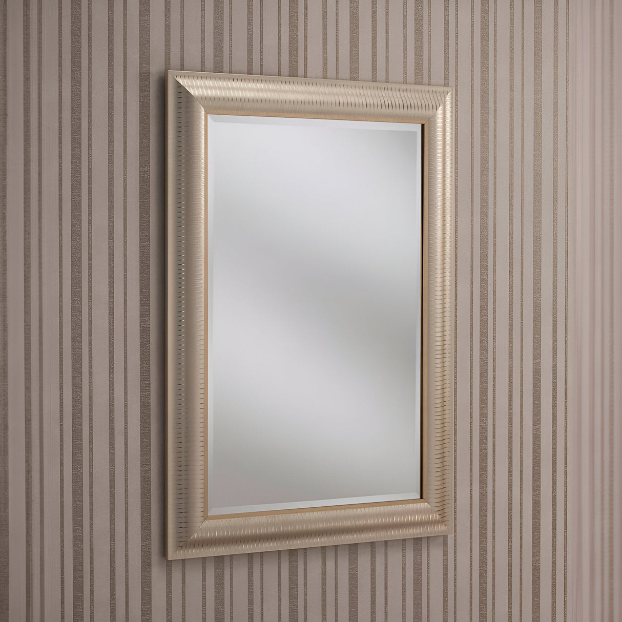 The Veris Rectangle Wall Mirror - Champagne, with its textured champagne-gold frame, is mounted on a wall decorated with vertical beige and brown striped wallpaper. The mirror reflects light beautifully, enhancing the modern design with its bright appearance.