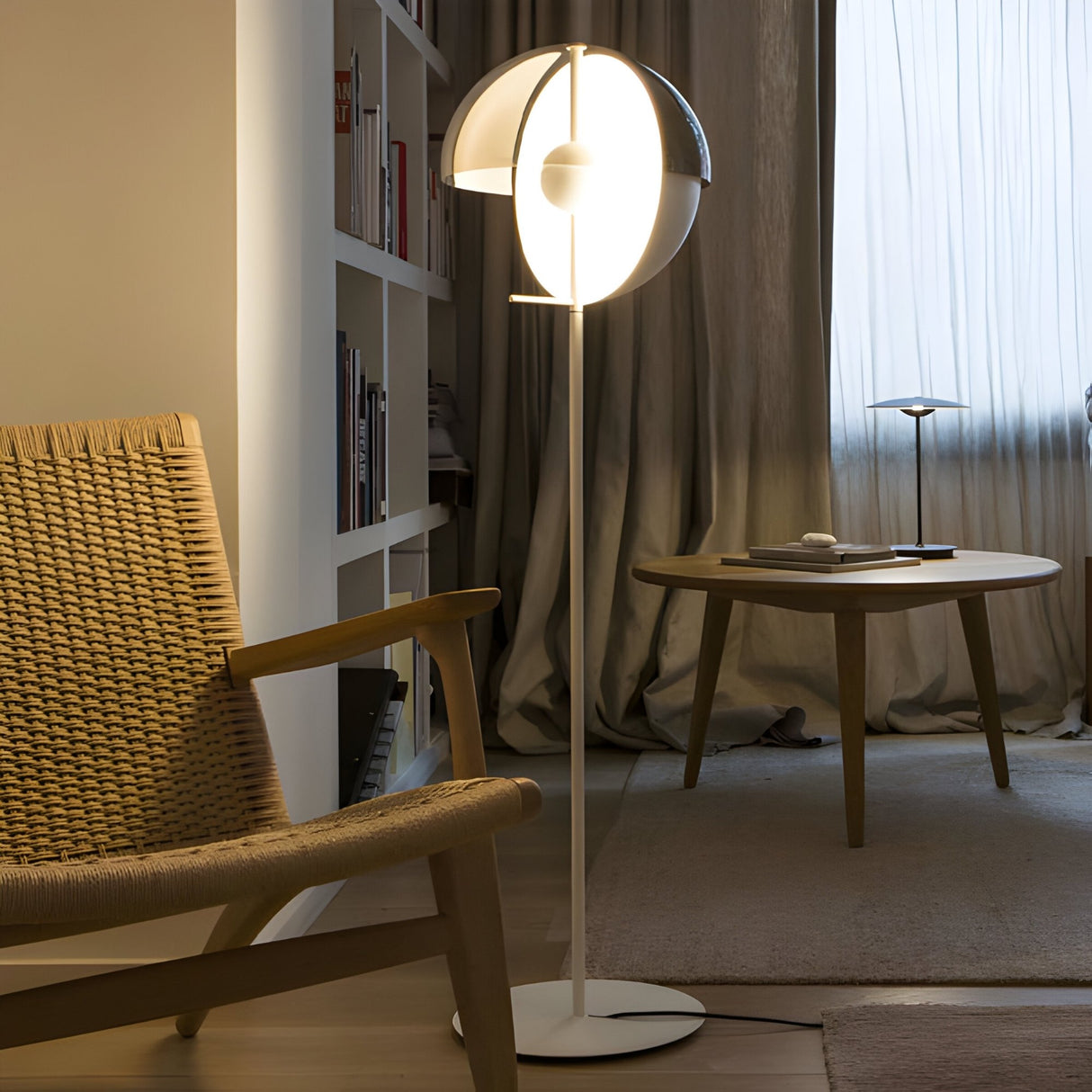 A modern living room with a woven armchair, round wooden table, bookshelves, and the Lucia LED Floor Lamp - White emitting warm light near sheer curtains covering a window.