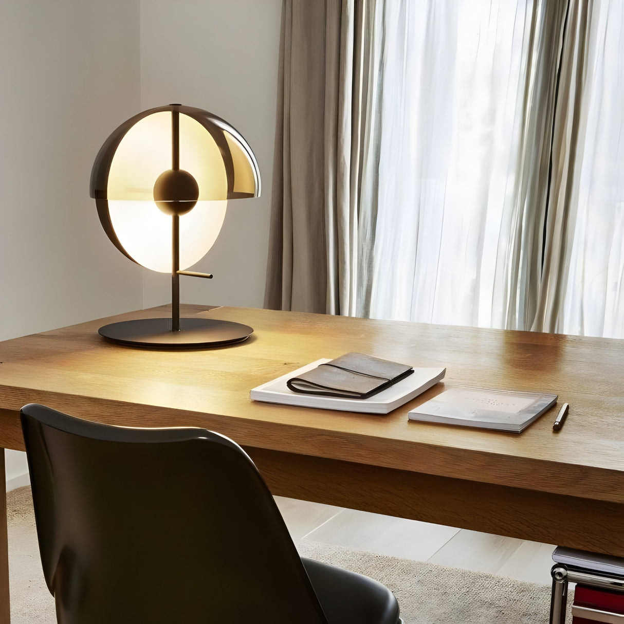 The Lucia LED Table Lamp - Black with a circular design sits on a wooden desk beside a notebook, closed book, and pen. A black chair is in front of the desk, and sheer curtains cover the window, providing energy-efficient lighting.