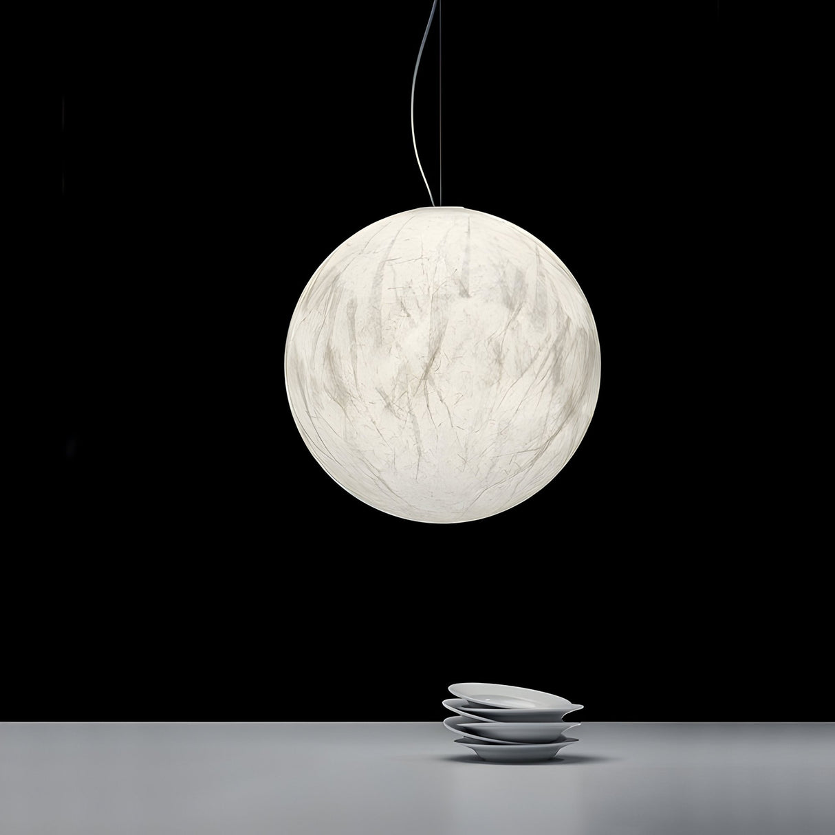 The large, round Lunar Pendant Light - White with a soft sheen hangs above a minimalistic surface with four stacked white plates, set against a black background.
