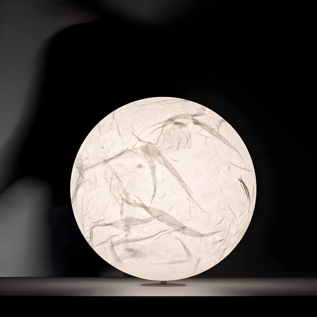 The Lunar Table Lamp - White features a spherical, glowing design with a textured, crumpled paper-like surface made from hand-painted silk-infused acrylic, and sits elegantly on a flat surface against a dark background.