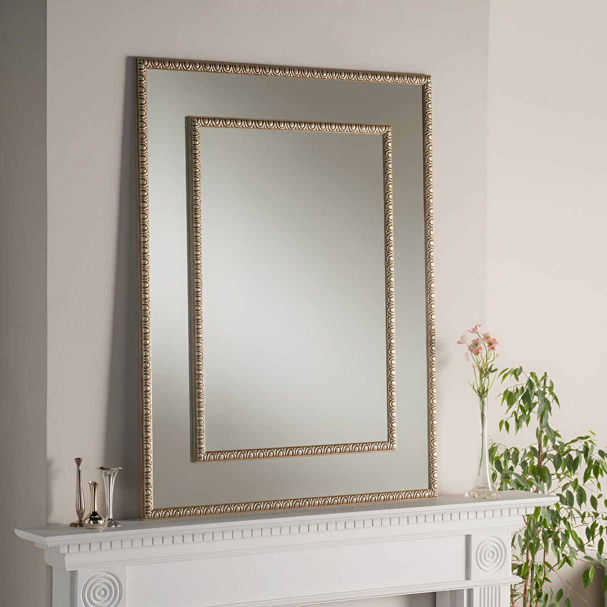 A Virelle Rectangle Wall Mirror - Champagne Gold graces the beige wall above a white mantel, where two silver vases accompany pink flowers in a glass vase. A carved wooden frame adds sophistication, and a leafy plant adorns the right side.