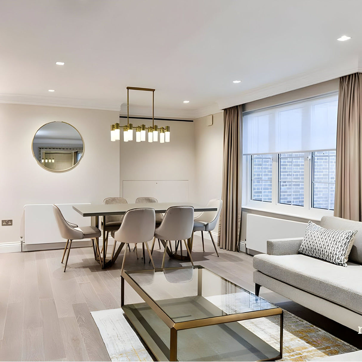 Modern dining and living room with a rectangular glass coffee table, grey sofa, dining table for six, Magari 10 Linear Suspension Light - Gold with opal shades, round wall mirror, large windows with beige curtains, and light wood flooring.