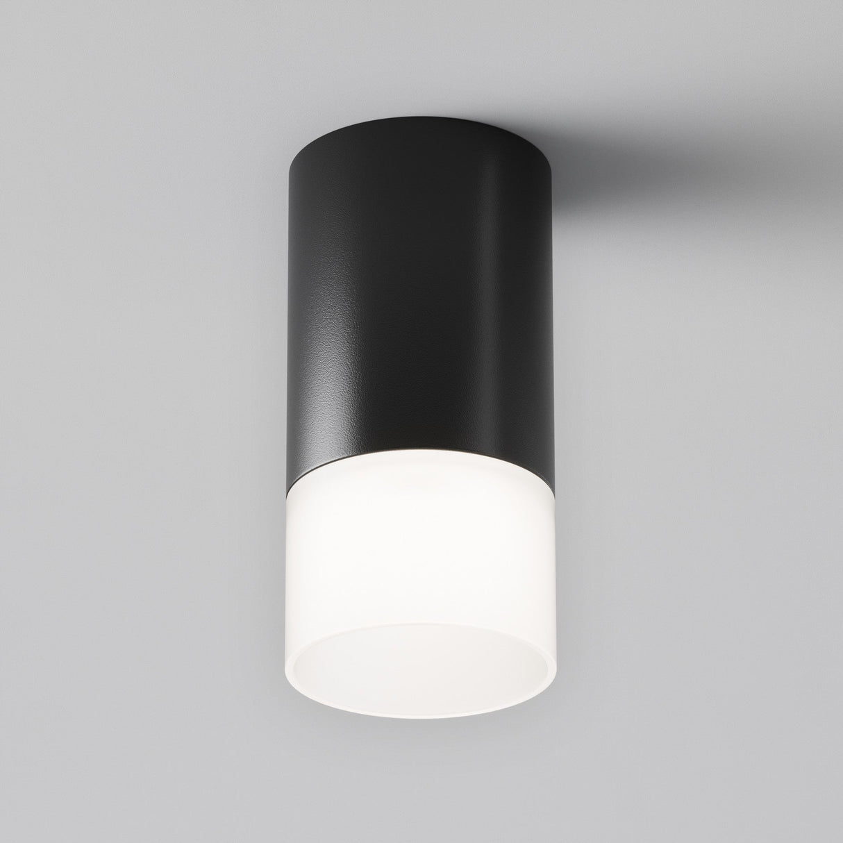 The Pauline Long Round Surface Downlight - Black showcases modern minimalism with its sleek design, a black top, and a frosted white diffuser, offering soft ambient lighting elegantly on a smooth light gray ceiling.