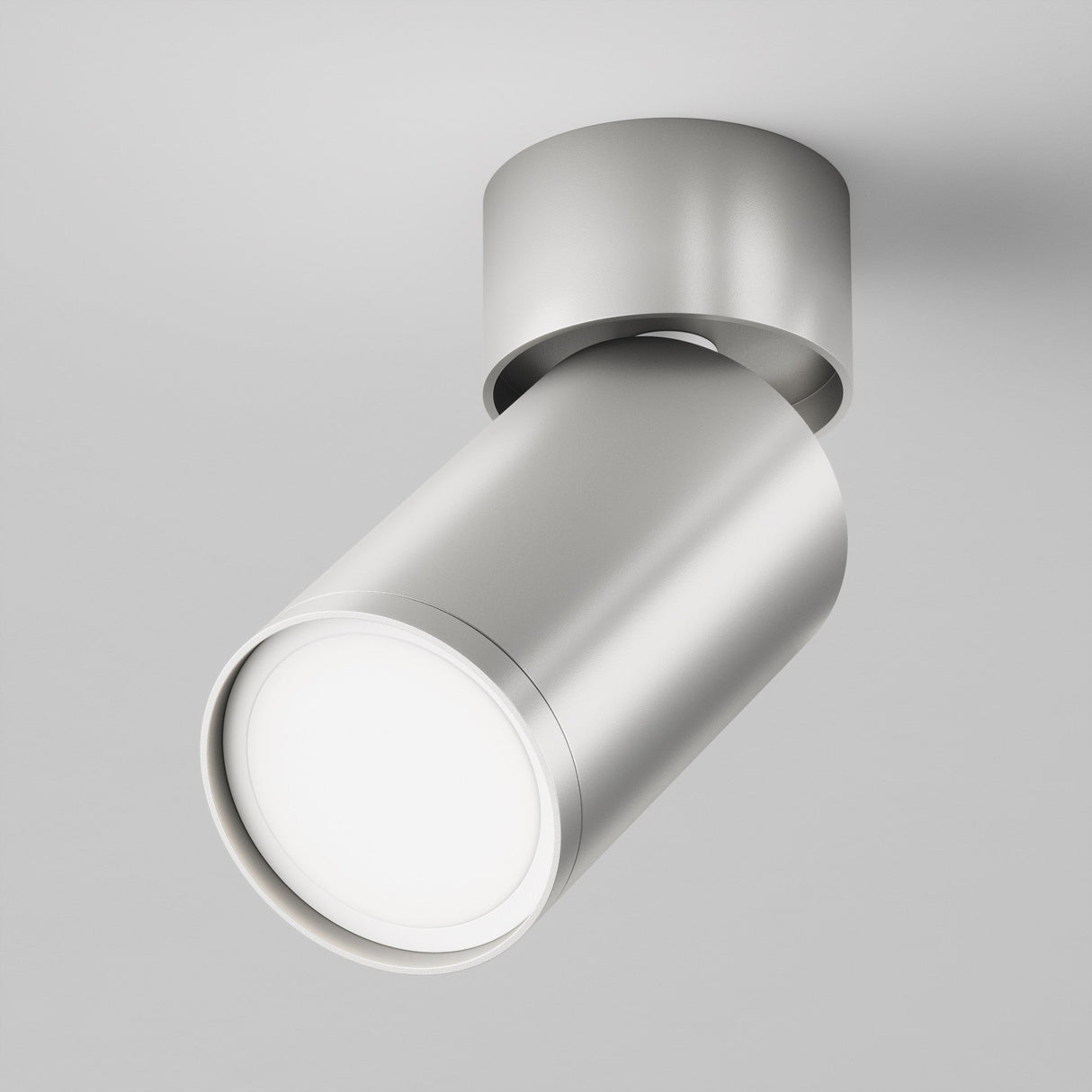 The Focus S Single Surface-Mounted Spotlight - Silver is a modern cylindrical light with a sleek finish, mounted on a round base and angled downward. It emits a bright, focused beam against a plain white wall.