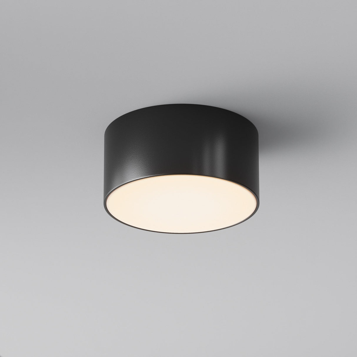 The Zon Outdoor Ceiling Light 15W 3000K - Black features a sleek, black cylindrical design and IP65 rating, mounted on a light gray ceiling. It emits warm, soft light, enhancing the minimalist aesthetic of any space.