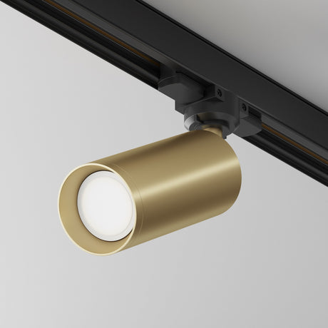 The Focus 3-Circuit Trinity Track Spotlight in matte gold features a sleek cylinder design on a black rail, ideal for enhancing industrial interiors with its modern appeal against a white backdrop.